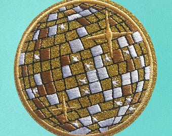 May include: A gold and white glitter embroidered iron-on patch of a disco ball. The patch is round and has a white border.