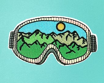 May include: Embroidered patch of a pair of ski goggles with a mountain landscape and a yellow sun in the lens. The mountains are green and brown, and the sky is blue.
