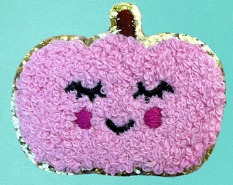 May include: A pink pumpkin-shaped patch with a textured, fuzzy surface. It features embroidered black closed eyes with long lashes, a small black smile, and two bright pink circular cheeks. The patch has a dark brown stem and a gold glitter border.