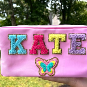 May include: Pink zippered pouch with the name "KATE" in colorful, fuzzy letters with gold glitter trim. A gold glitter butterfly with turquoise and yellow accents is centered below the name.