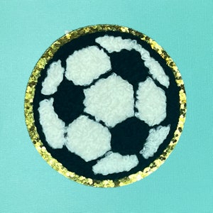 May include: A white and black soccer ball with a gold glitter border. The ball is made of a fuzzy material.