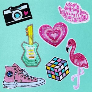 May include: A set of nine embroidered iron-on patches featuring a pink heart, a pink flamingo, a pink cowboy hat, a green electric guitar, a pink and white Rubik's cube, a pink and white camera, a pink and white sneaker, and a pink heart with a red heart inside.
