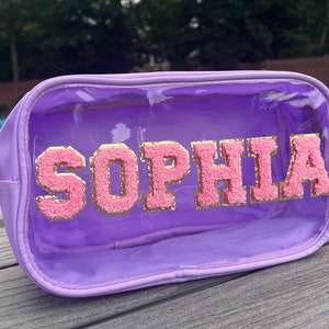 May include: A clear purple cosmetic bag with a zipper closure. The bag has a pink and gold glitter name "Sophia" on the front.
