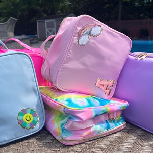 May include: Four colorful zippered pouches with different embroidered designs. The pouches are light blue, pink, purple, and tie-dye. Each pouch has a gold letter 'A' and a different design, including a rainbow, a unicorn, a flower, and a tie-dye pattern.