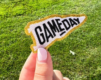 May include: A white felt pennant with gold glitter trim and the text "GAMEDAY" in black embroidery.