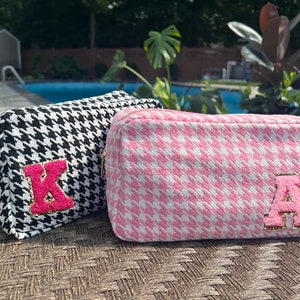 May include: Two zippered cosmetic bags with a houndstooth pattern. One bag is black and white, with a pink letter "K" on the front. The other bag is pink and white, with a pink letter "A" on the front.