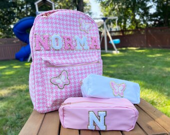 May include: Pink and white houndstooth backpack with the name "NORMA" in gold letters and butterfly embellishments.  Two matching pink and white pencil cases with butterfly and letter embellishments.