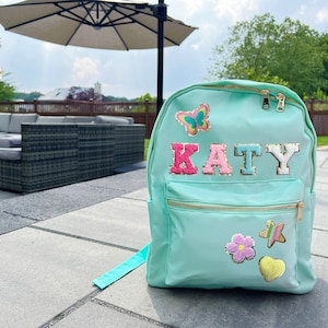 May include: A light blue backpack with a gold zipper and a gold star, flower, and heart patch. The backpack has the name "KATY" spelled out in colorful letters.