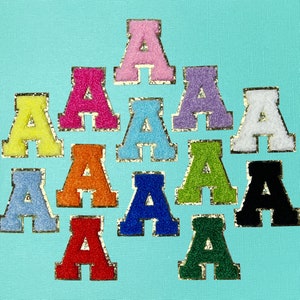 May include: A collection of colorful, embroidered letter "A" patches. Each letter is a different color, including pink, yellow, orange, blue, green, and black, with a gold glitter border. The patches are arranged on a light blue background.