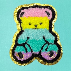 May include: A colorful fuzzy bear patch with a gold glitter border. The bear is pink, yellow, blue, and green.