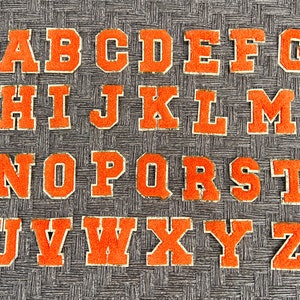 May include: Orange felt letters of the alphabet, A through Z, on a gray textured background.
