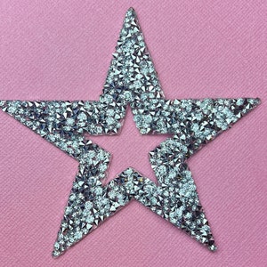 May include: A sparkling star-shaped embellishment, covered in numerous small, reflective rhinestones. The star is set against a soft pink background, creating a contrast that highlights the star's glittery texture. This decorative element could be used for crafts or fashion.