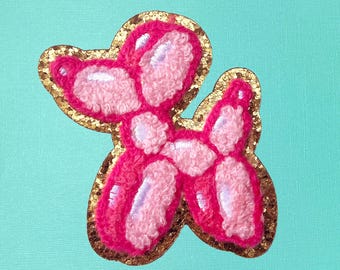 May include: A pink and gold balloon animal patch. The balloon animal is shaped like a dog. The patch is made of felt and has a gold glitter outline.