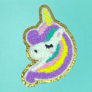 May include: A purple, pink, yellow, and green unicorn applique with a gold glitter outline. The unicorn has a closed eye and a yellow horn.