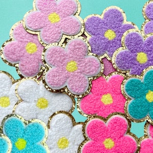 May include: A collection of colorful fuzzy flower patches with gold glitter trim. The flowers are in shades of pink, purple, blue, and white.