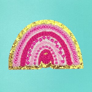 May include: A pink and white rainbow embroidered patch with a gold glitter border. The rainbow is made of yarn and has a layered effect.
