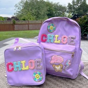 May include: Purple backpack and lunch bag set with the name "CHLOE" in colorful, fluffy letters. Both bags feature embroidered patches of a flower, butterfly, cloud, and lightning bolt.