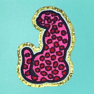 May include: A pink leopard print patch with black and red spots, outlined in black and white, and edged with gold glitter. The patch is set against a light teal background.