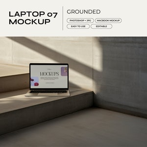 Op de afbeelding: Een zilveren laptop met een zwart scherm waarop het woord "Mockups" wordt weergegeven, op een lichtbruin houten oppervlak. De laptop staat op een trap in een moderne betonnen ruimte.