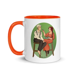 May include: White ceramic mug with an orange rim, featuring an illustration of two people sitting at a table, sharing a meal. The man has short blonde hair and is wearing a white shirt with a brown vest. The woman has long red hair and is wearing a red dress. The background is a green circle.