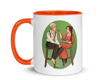 May include: White ceramic mug with an orange rim, featuring an illustration of two people sitting at a table, sharing a meal. The man has short blonde hair and is wearing a white shirt with a brown vest. The woman has long red hair and is wearing a red dress. The background is a green circle.