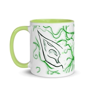May include: White ceramic mug with a green and white design. The design features a stylized ear with a green outline and a white interior. The mug has a light green rim.