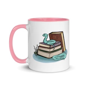 May include: A white ceramic mug with a pink rim features a whimsical illustration of a blue snake curled around a stack of books. The books are brown, purple, and blue with a brown cover on top.