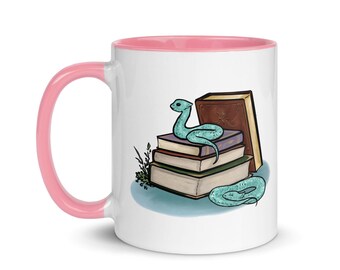 May include: A white ceramic mug with a pink rim features a whimsical illustration of a blue snake curled around a stack of books. The books are brown, purple, and blue with a brown cover on top.