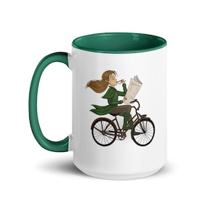 May include: White ceramic mug with a green rim. The mug features a cartoon illustration of a person riding a bicycle while reading a newspaper and holding a cup of coffee.