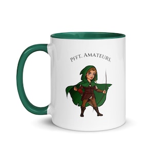 May include: White ceramic mug with a green interior and handle. The mug features a cartoon illustration of a woman in a green hooded cloak holding a bow and arrow. Text above the illustration reads "Pfft. Amateurs."