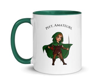 May include: White ceramic mug with a green interior and handle. The mug features a cartoon illustration of a woman in a green hooded cloak holding a bow and arrow. Text above the illustration reads "Pfft. Amateurs."