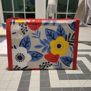 May include: A small zippered pouch with a red border and a white background featuring a floral pattern with blue leaves, yellow flowers, and red berries.
