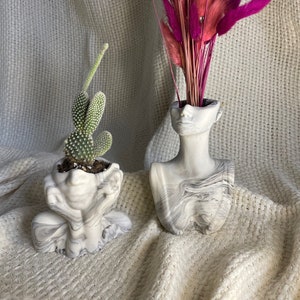 Peut inclure: Deux pots en céramique à motif marbré blanc, l'un contenant un cactus et l'autre un bouquet de fleurs séchées roses.