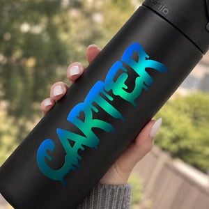 May include: A matte black water bottle featuring the name CARTER in a vibrant, iridescent blue-to-green gradient. The lettering has a distinct dripping paint effect, with the text oriented vertically along the side of the cylindrical container.