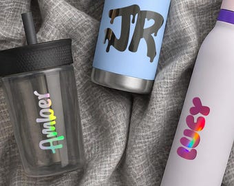 Puede incluir: Un vaso de plástico transparente con tapa y pajita negra con el nombre Amber en letras iridiscentes, una botella de metal azul claro con las letras negras goteantes JR y una botella blanca con el nombre Lucy en degradado rosa.