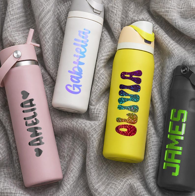 Puede incluir: Cuatro botellas de agua cil&iacute;ndricas con tapas y nombres personalizados. Una botella rosa claro con el nombre AMELIA, una blanca con Gabriella, una amarilla con Olivia en texto arco&iacute;ris brillante y una negra con JAMES.