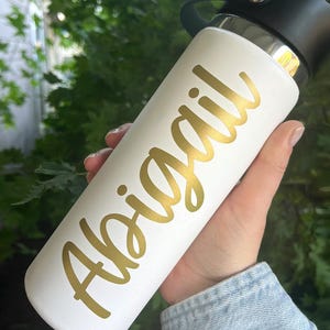 May include: White insulated water bottle with a black lid. The name "Abigail" is printed in gold script on the side. The bottle is held against a background of green foliage.
