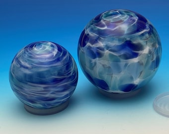 May include: Two decorative glass spheres with blue and white swirling patterns. The spheres sit on grey bases, with one sphere larger than the other. The glass has a textured appearance. The background is a blue gradient.