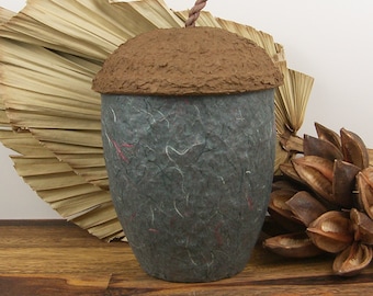 Op de afbeelding: Een handgemaakte, eikelvormige container met een getextureerde, donkergrijze body en een bruin, getextureerd deksel. Het deksel heeft een gedraaide, bruine steel. De container staat op een houten oppervlak, met gedroogde bladeren en zaaddozen op de achtergrond.