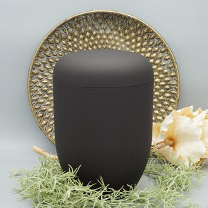 Op de afbeelding: Een zwarte matte urn met een afgeronde bovenkant en een glad oppervlak. De urn staat op een decoratief gouden bord.