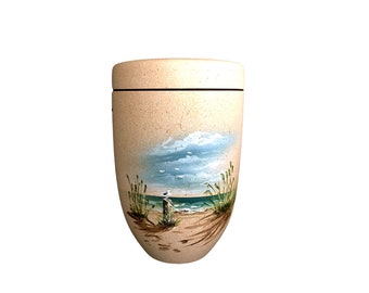 Op de afbeelding: Een beige urn met een handgeschilderde strandscène. Het kunstwerk toont een blauwe oceaan, een zandstrand en een zeemeeuw op een houten paal onder een bewolkte hemel. De urn heeft een zwarte band rond de bovenkant en een deksel.