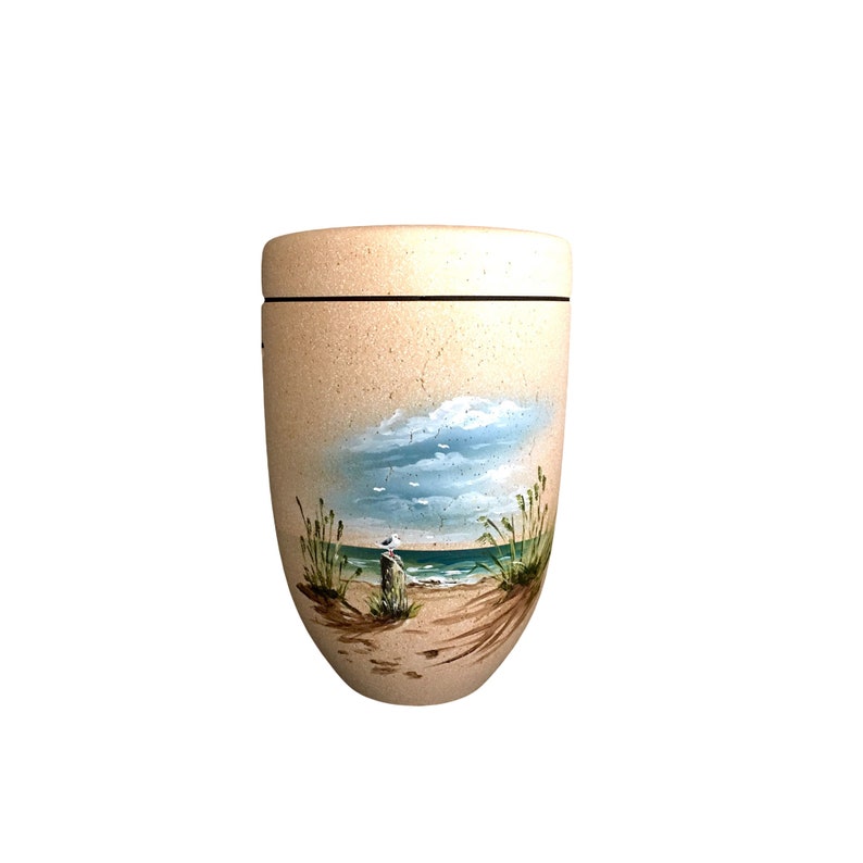 May include: A beige urn with a hand-painted beach scene. The artwork depicts a blue ocean, sandy shore, and a seagull perched on a wooden post under a cloudy sky. The urn has a black band around the top and a lid.