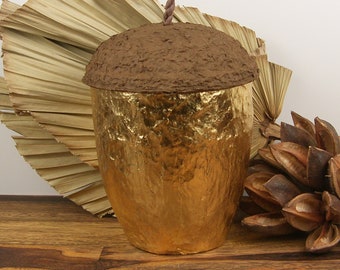 May include: A decorative acorn-shaped container with a textured brown top and a gold-colored body. The container has a twisted brown stem and sits on a wooden surface. Dried leaves and seed pods are in the background.