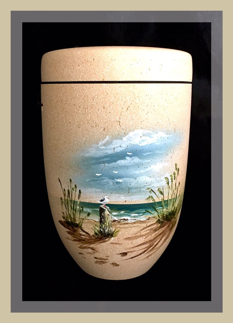 May include: A beige, urn-shaped container with a painted beach scene. The artwork depicts a blue sky with clouds, a sandy beach, ocean waves, and a seagull perched on a wooden post. The container has a black band around the top.