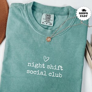 Puede incluir: Una camiseta de cuello redondo en color verde salvia con un estampado gráfico blanco que dice night shift social club con un pequeño icono de corazón sobre el texto. La prenda incluye una etiqueta de la marca Comfort Colors y un collar dorado.