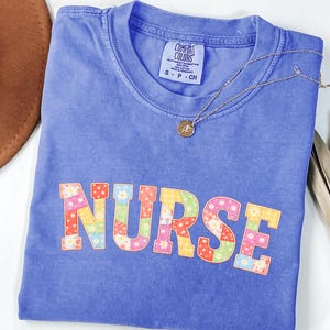 Puede incluir: Una camiseta azul Comfort Colors con la palabra "NURSE" en letras de patchwork coloridas. Un collar de oro con un colgante está drapeado sobre la camisa. La camisa está doblada sobre una superficie neutra.