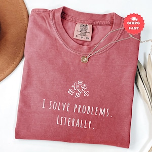 Pode incluir: Uma camiseta rosa empoeirada com o texto "I SOLVE PROBLEMS. LITERALLY." em branco. Acima do texto, há símbolos matemáticos. Um colar de prata com um pingente está sobre a camiseta. A camiseta tem gola redonda e mangas curtas.