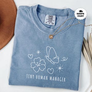 Può includere: Una maglietta blu Comfort Colors con una grafica bianca di una farfalla, un fiore, cuori e la scritta "TINY HUMAN MANAGER". Una collana d'oro con un ciondolo è drappeggiata sulla maglietta.