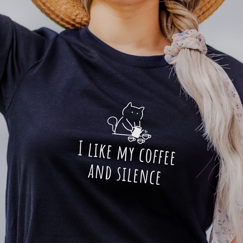 May include: A navy blue t-shirt with the text "I LIKE MY COFFEE AND SILENCE" in white. Above the text is a white line drawing of a cat holding a coffee cup. The shirt is a casual style.