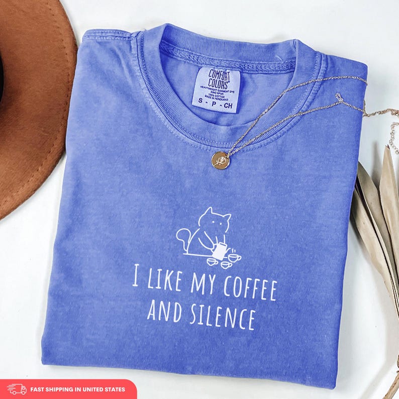 May include: A blue Comfort Colors t-shirt with the text "I LIKE MY COFFEE AND SILENCE" and a cat graphic. A gold necklace with a pendant is draped across the shirt. The shirt is folded on a white surface.
